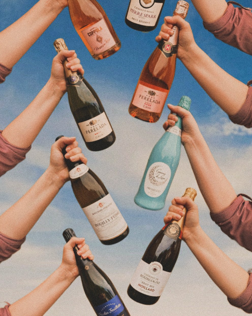 Eight bottles of champagne being held in the air all together in a line with the blue sky and a few clouds in the background.