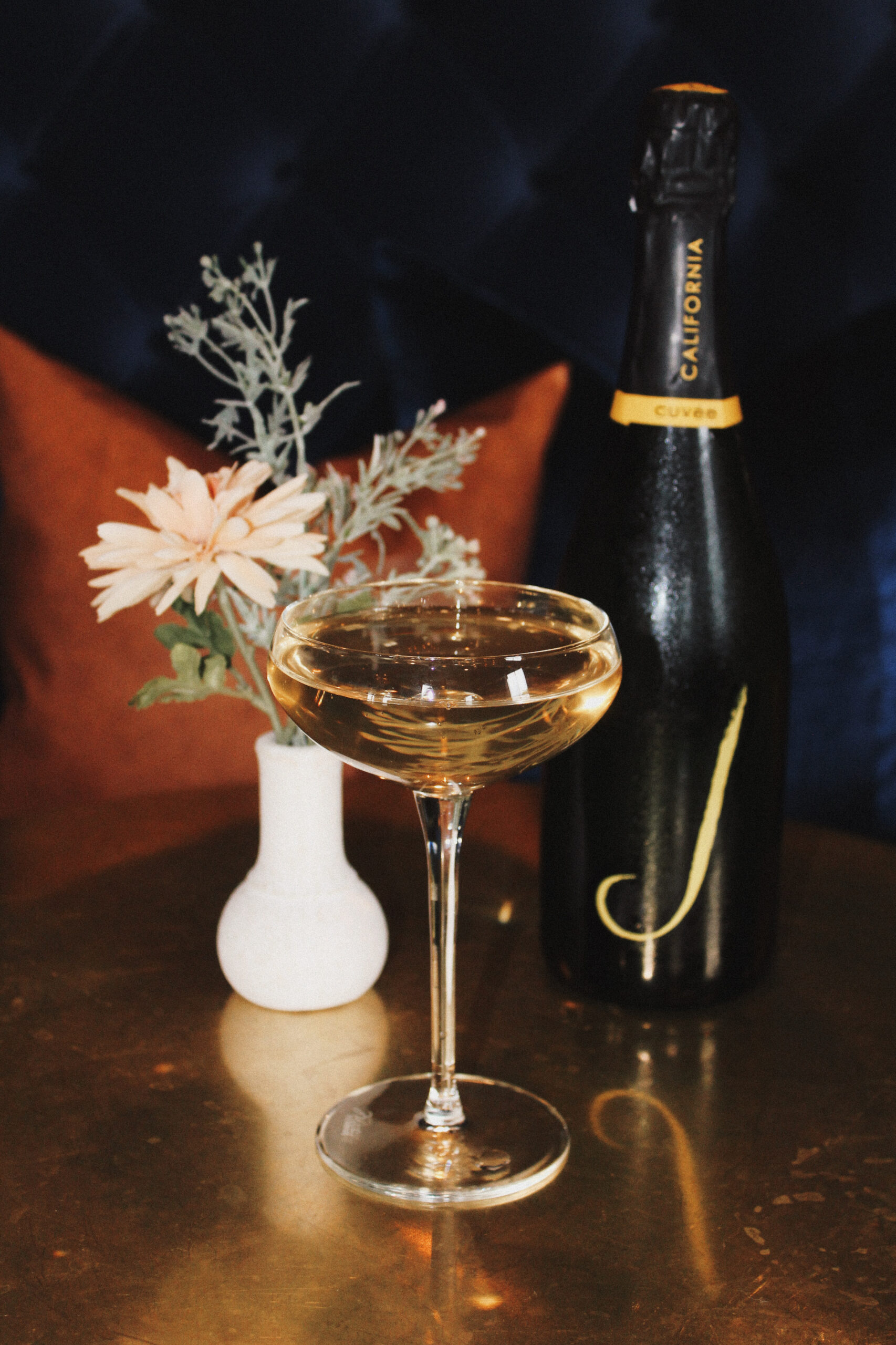 Gold table with a bottle of champagne and coup glass filled with bubbly. Vase of flowers and rust colored pillow in the background.